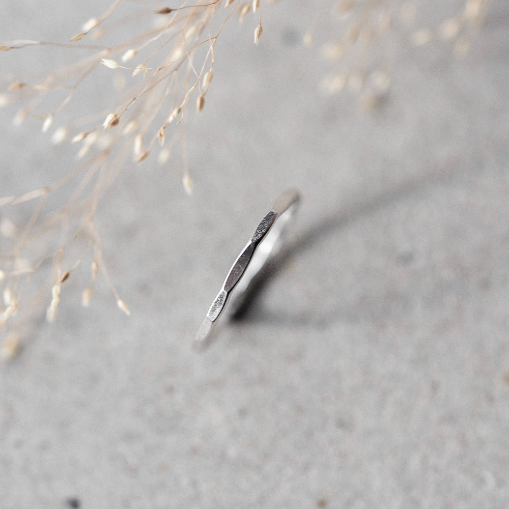 A simple, elegant silver stacking ring with a hammered finish, displayed on a grey background with dried plants beside it.