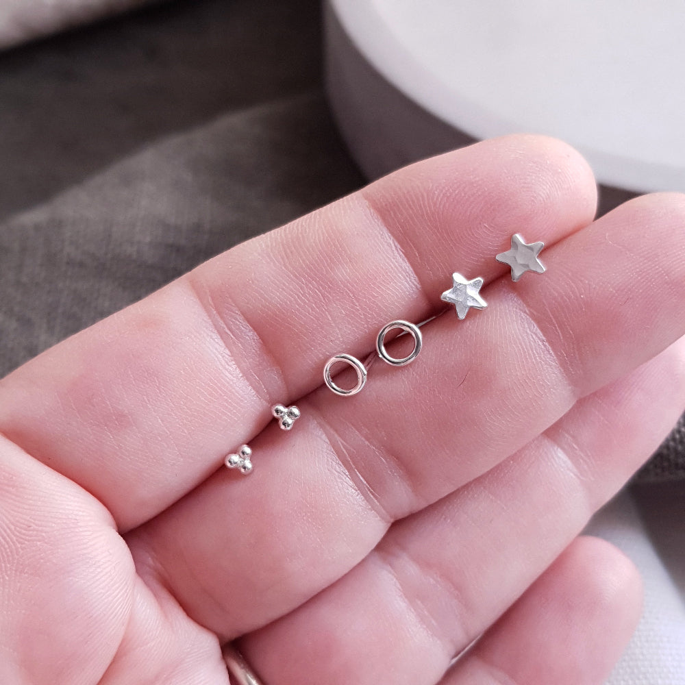 Set of 3 pairs of small silver stud earrings held between fingers. Set 1 is 3 small balls forming a triangle, set 2 are circles and set 3 are stars