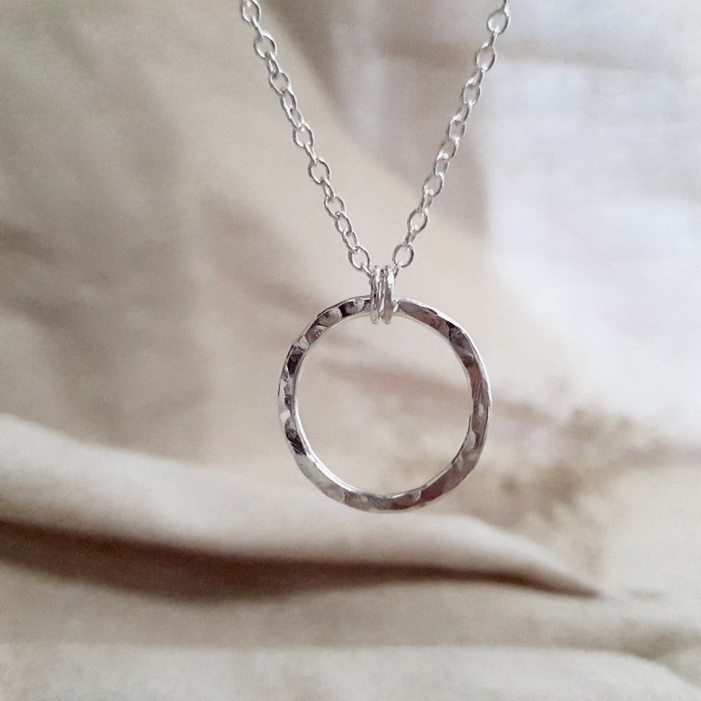 A silver eternity necklace with a hammered circle pendant and two little links connecting it to the chain, displayed on a beige fabric surface.
