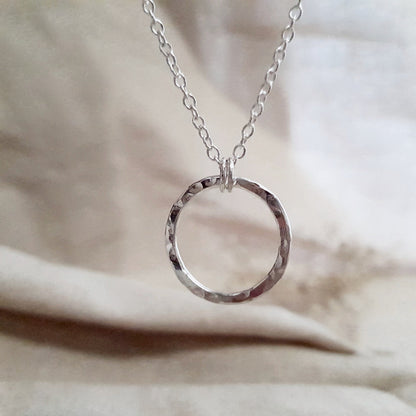 A silver eternity necklace with a hammered circle pendant and two little links connecting it to the chain, displayed on a beige fabric surface.