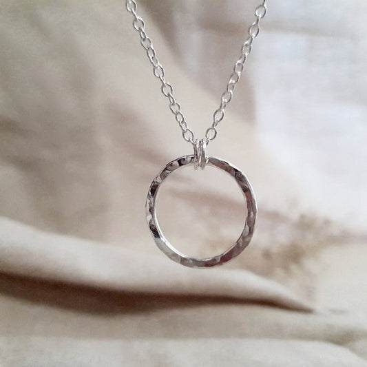 A silver eternity necklace with a hammered circle pendant and two little links connecting it to the chain, displayed on a beige fabric surface.