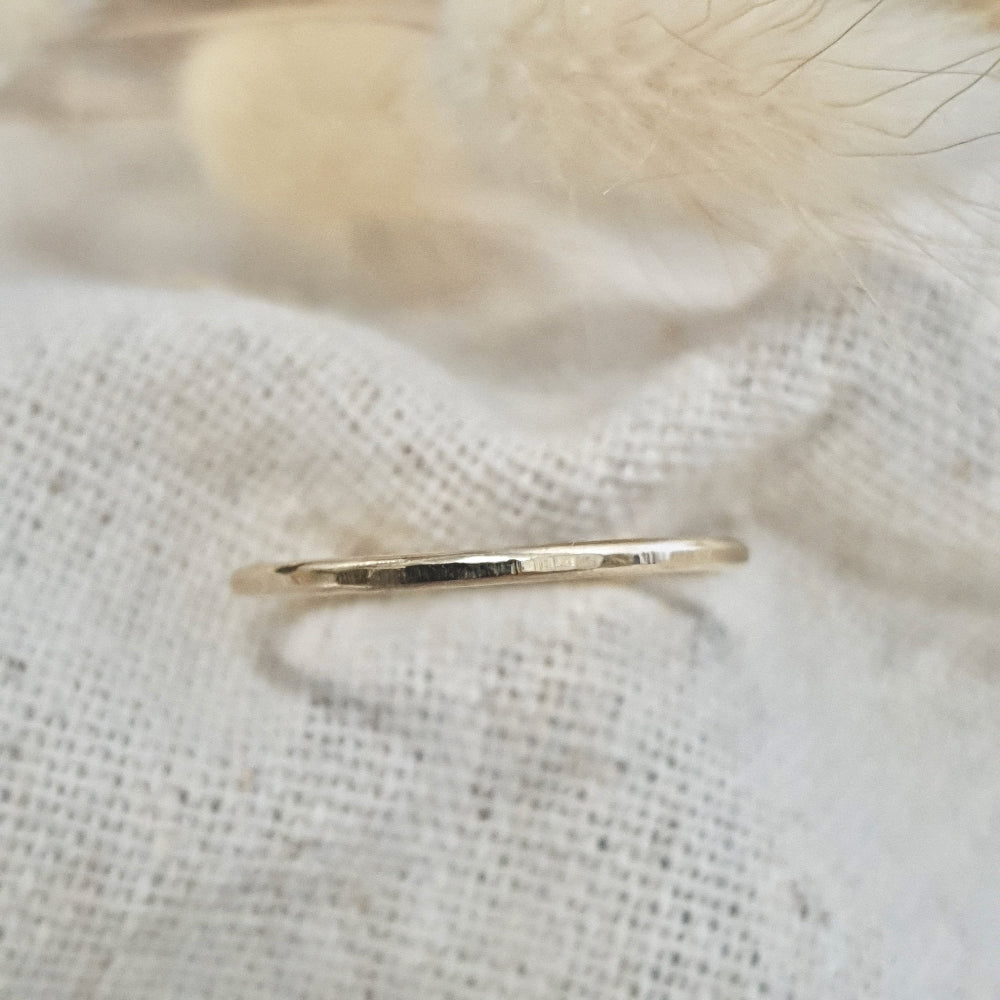 A flat textured 9ct gold stacking ring on a linen surface, with a hammered finish and  on white linen with a feather behind it.