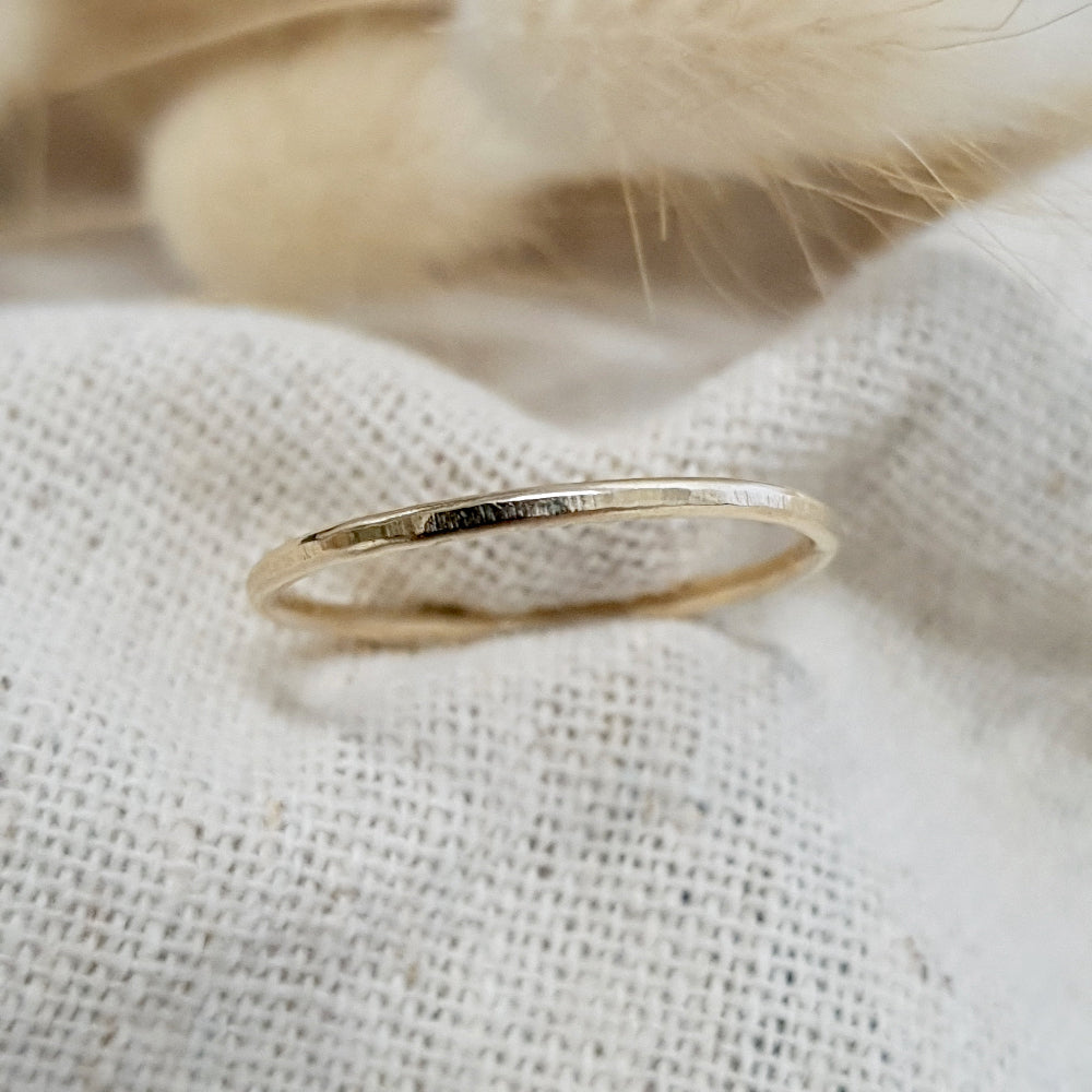 A flat textured 9ct gold stacking ring on a linen surface, with a hammered finish and  on white linen with a feather behind it.