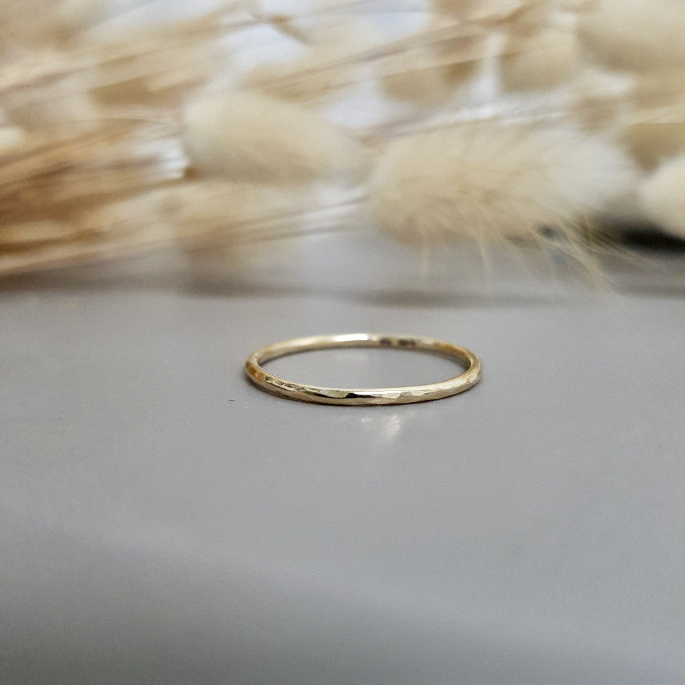 A single hammered gold stacking ring displayed against a neutral background with dried flowers to the side.