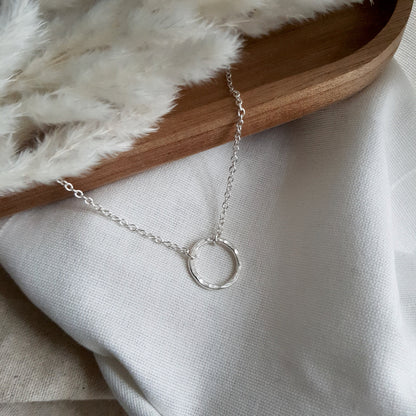 hammered Silver circle pendant with chain attached each side on a white fabric background with a wooden tray and white dried grasses