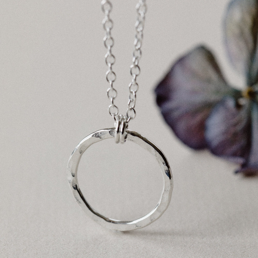 A silver eternity necklace with a hammered circle pendant and two little links connecting it to the chain, on a white background with a blurred purple flower 