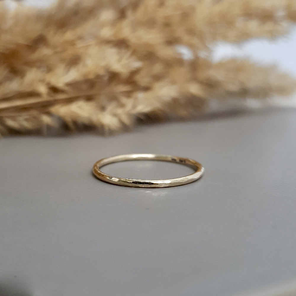 A single hammered gold stacking ring displayed against a neutral background with dried flowers to the side.