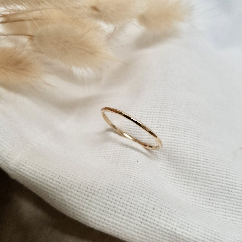 A single hammered gold stacking ring displayed against a neutral background with dried flowers to the side.