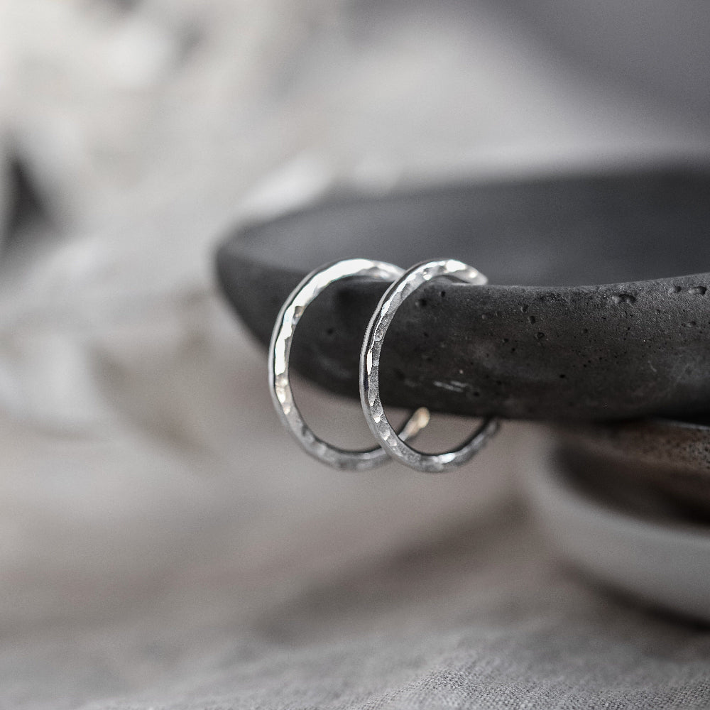 A pair of sterling silver hammered hoop earrings resting on the edge of a dark grey stone dish with a white blurred background
