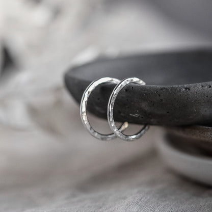 A pair of sterling silver hammered hoop earrings resting on the edge of a dark grey stone dish with a white blurred background