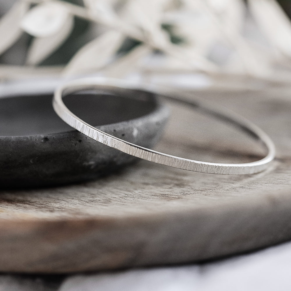 A silver bangle bracelet with a hammered texture, displayed on a wooden surface.