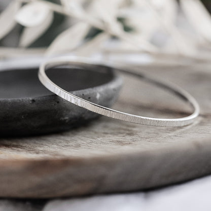 A silver bangle bracelet with a hammered texture, displayed on a wooden surface.