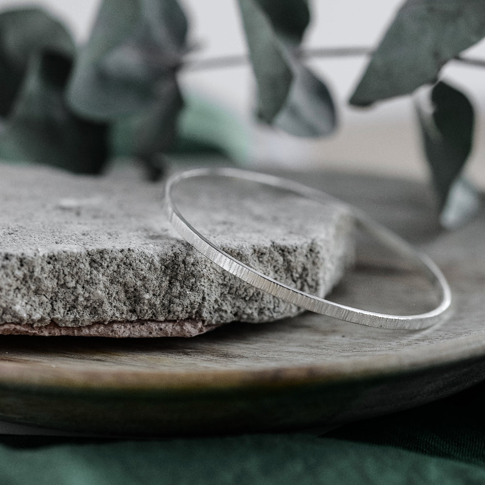 A silver bangle bracelet with a hammered texture, displayed on a stone surface.