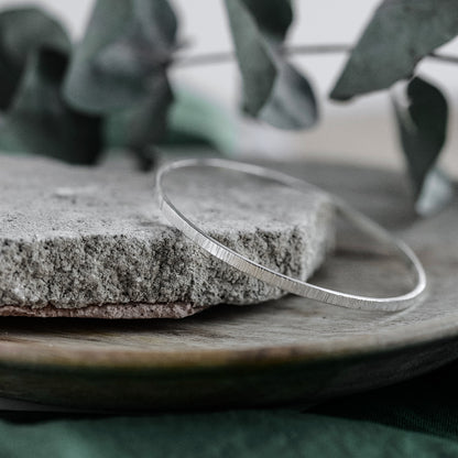 A silver bangle bracelet with a hammered texture, displayed on a stone surface.