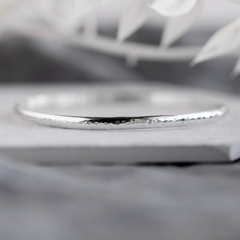 A silver bangle bracelet with a hammered texture, displayed on a grey surface.