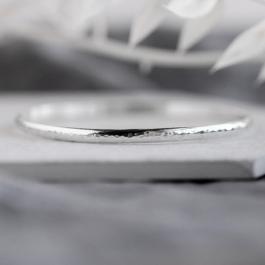 A silver bangle bracelet with a hammered texture, displayed on a grey surface.