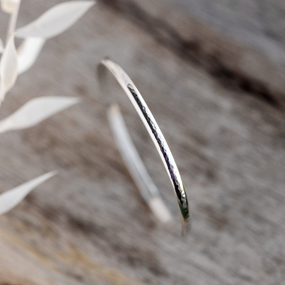 A silver bangle bracelet with a hammered texture, displayed on a pale wooden surface.