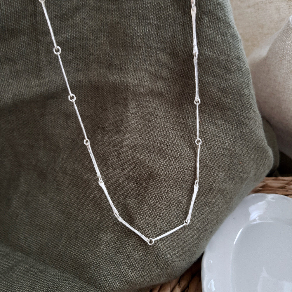 A silver bar chain necklace with a hammered texture, displayed on a green textured background.