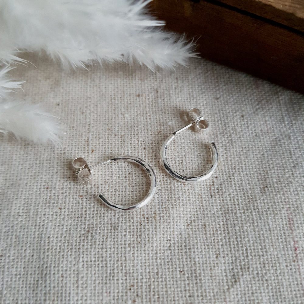 A pair of silver hammered hoop earrings resting on a cream textured fabric with a furry object in the background.