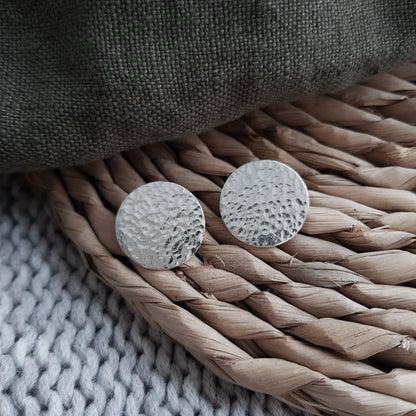 A pair of silver hammered disc earrings resting on a woven  surface.