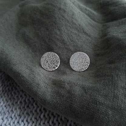 A pair of silver hammered disc earrings resting on a green fabric