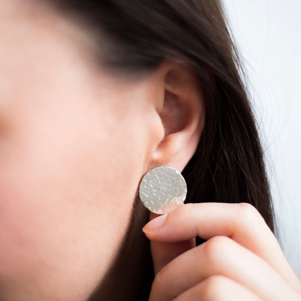 Large silver hammered disc earrings being worn by a female with long dark hair and she is touching the earring with her hand