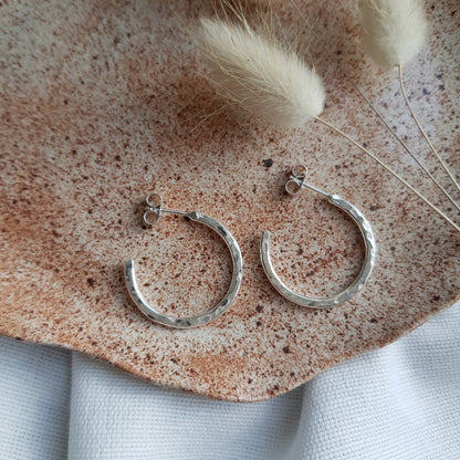 A pair of sterling silver hammered hoop earrings resting on a brown speckled surface with a fluffy white plant in the background.