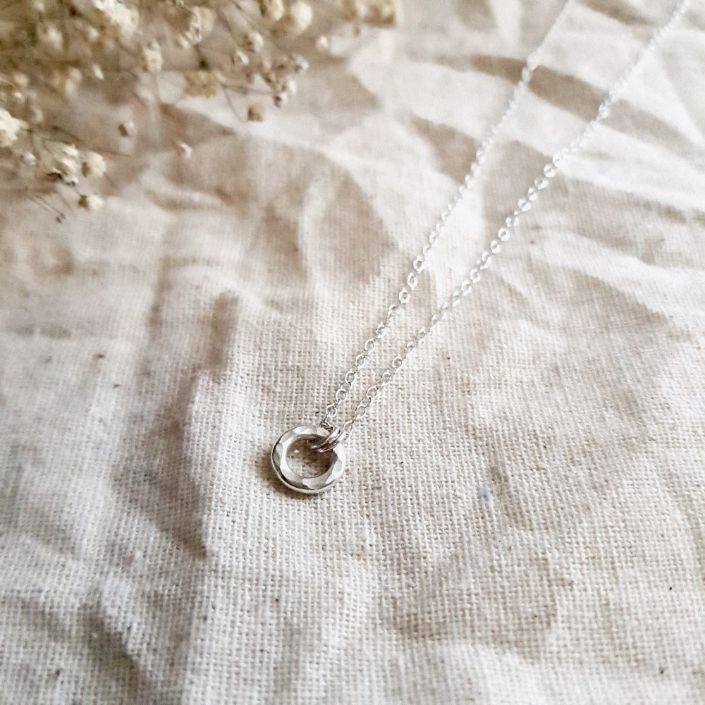 A sterling silver dainty necklace with a small hammered circular pendant, displayed on a cream fabric background.