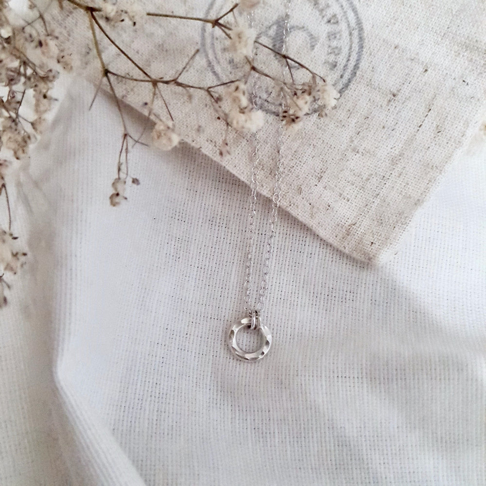 A sterling silver necklace with a small hammered circular pendant, displayed on a white fabric background.