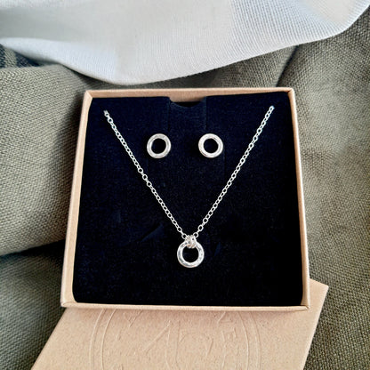 A silver dainty hammered circle necklace with matching stud earrings displayed in a brown craft box with a black insert