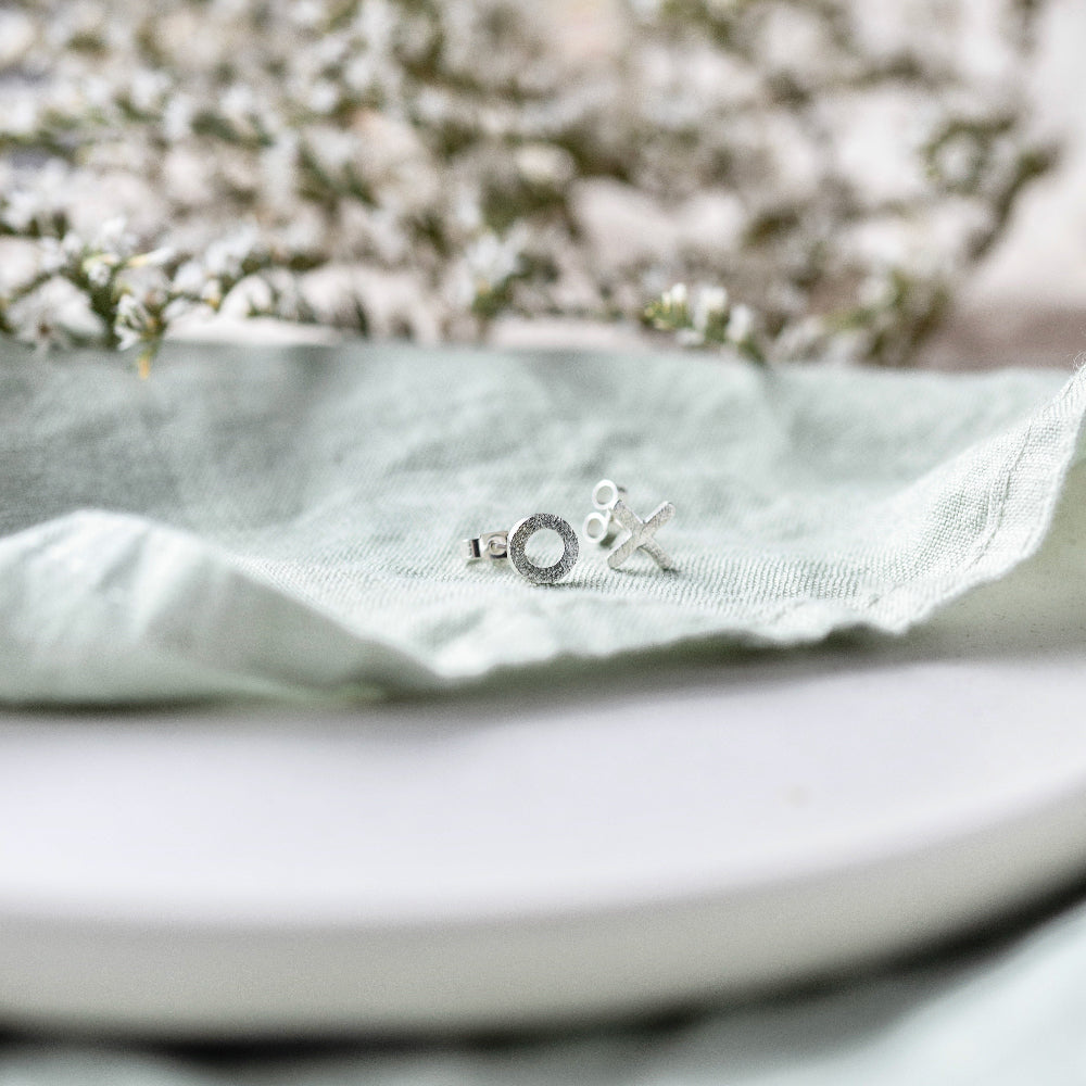 Hugs and Kisses silver stud earrings on a pale mint green fabric with foliage in the background