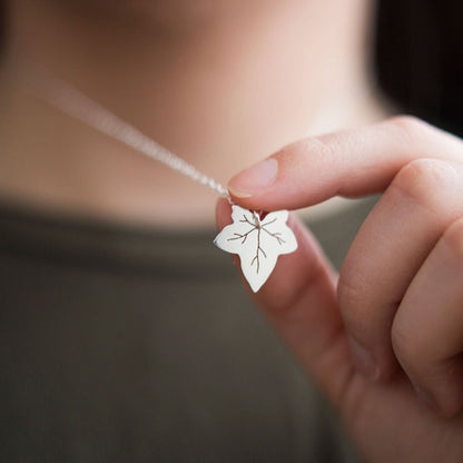 a lady  wearing a khaki green  T-shirt and a silver ivy leaf necklace she is holding the pendant between her thumb and first finger