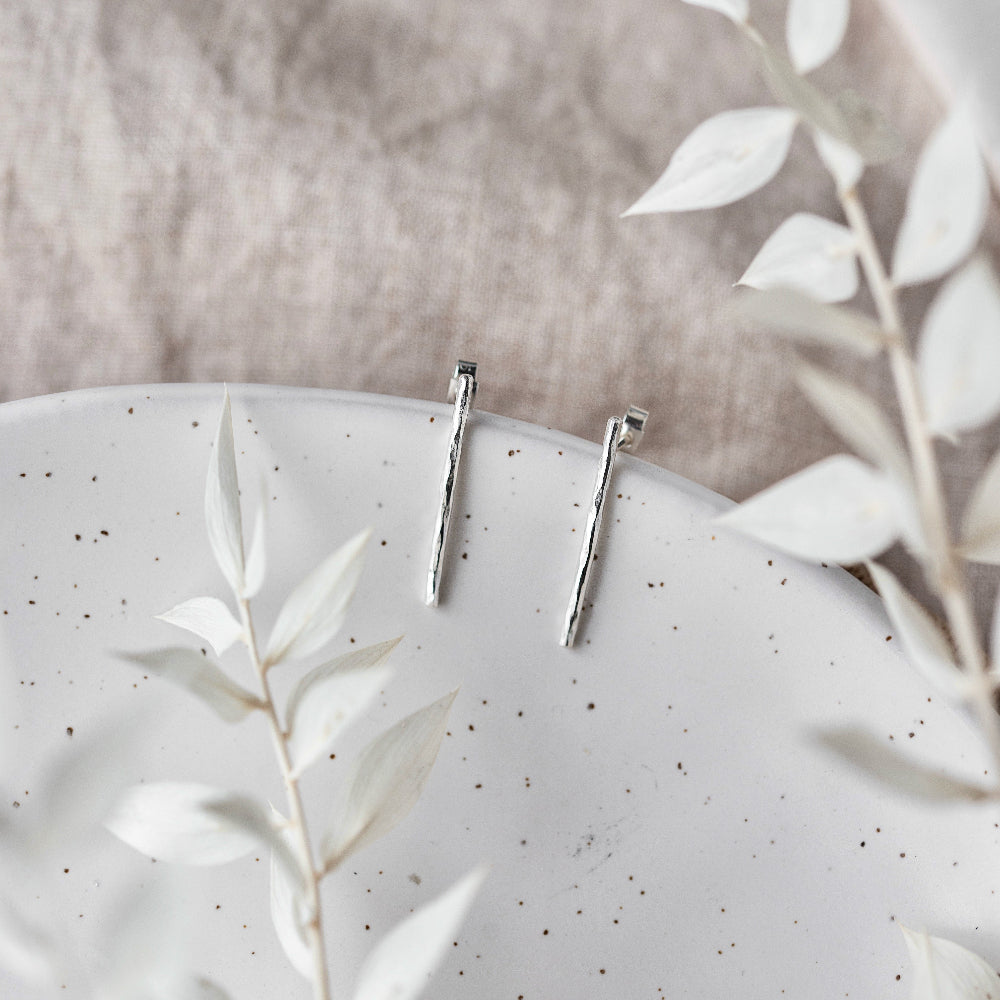 A pair of hammered silver bar earrings hanging from a cream ceramic dish with a blurred background and white leaves