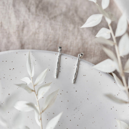 A pair of hammered silver bar earrings hanging from a cream ceramic dish with a blurred background and white leaves
