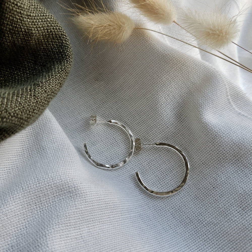 A pair of sterling silver hammered hoop earrings resting on a beige fabric surface with a fluffy white plant in the background.