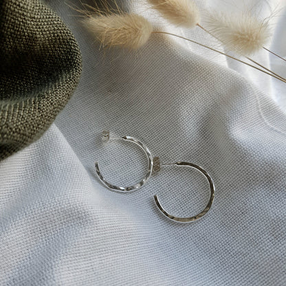 A pair of sterling silver hammered hoop earrings resting on a beige fabric surface with a fluffy white plant in the background.