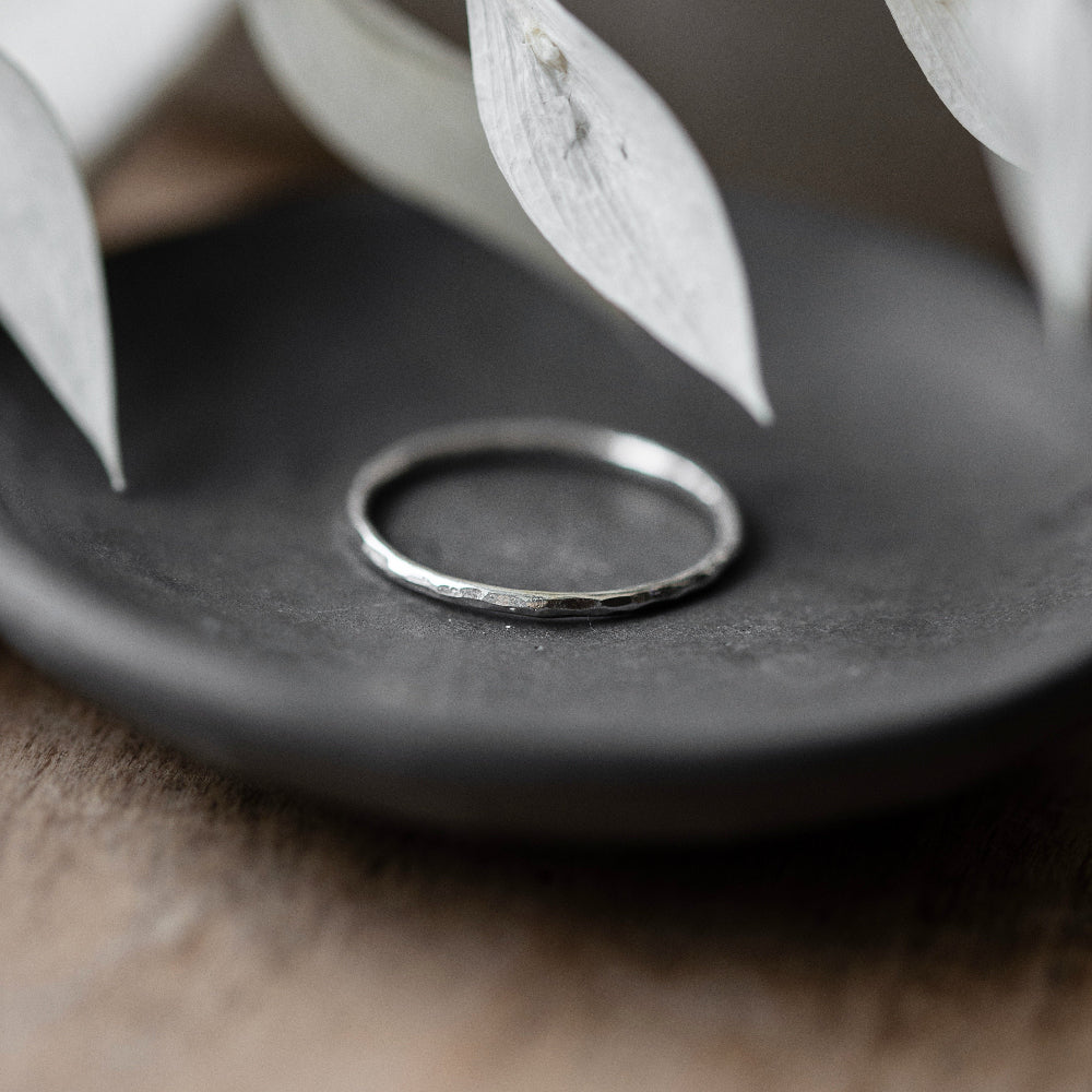 A single hammered silver stacking ring placed on a grey surface, with delicate white flowers hanging over it.