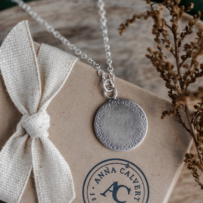 A silver necklace with a hammered texture pendant, displayed on a card with 'Anna Calvert' brand logp.