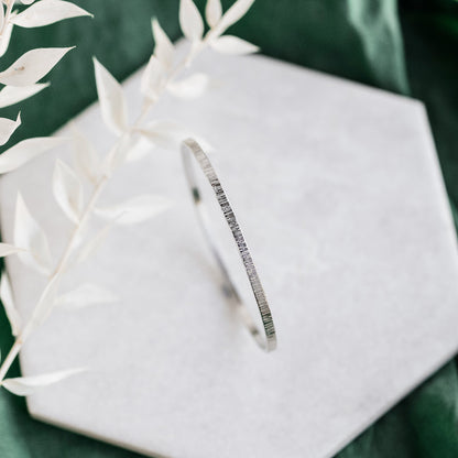 A silver bangle with a hammered lines texture, displayed on a white ceramic tile on a green background