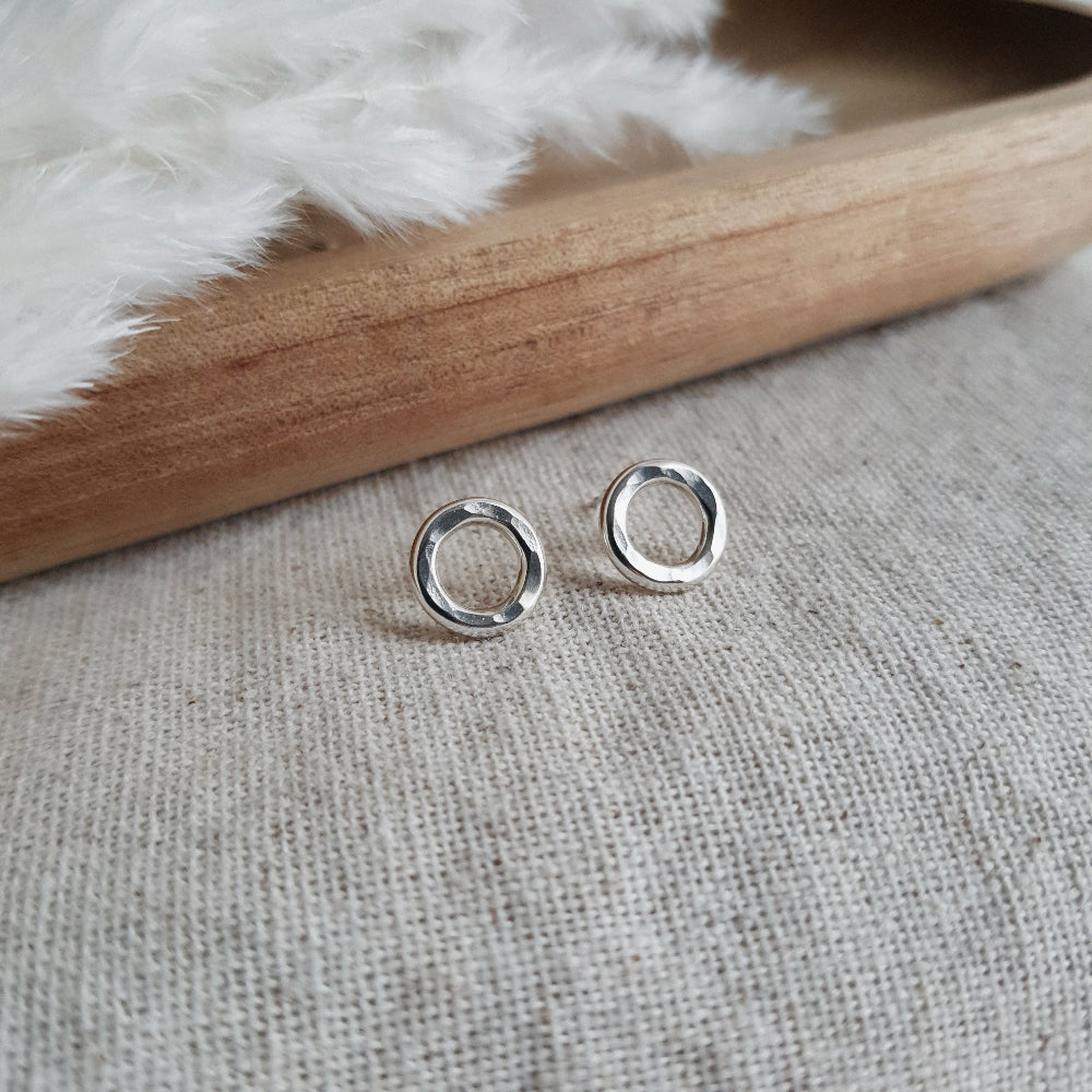 A pair of sterling silver hammered circle stud earrings resting on a white fabric surface with a wooden tray in the background