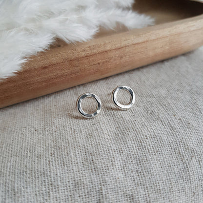 A pair of sterling silver hammered circle stud earrings resting on a white fabric surface with a wooden tray in the background