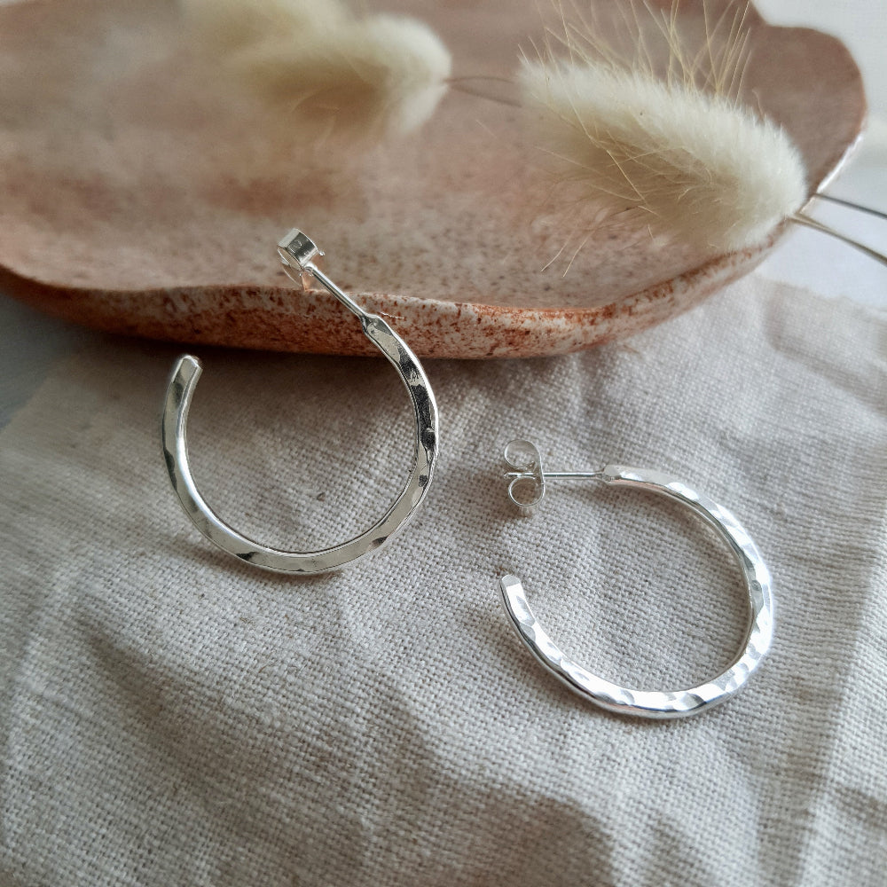A pair of sterling silver hammered hoop earrings resting on a beige fabric surface with a fluffy white plant in the background.