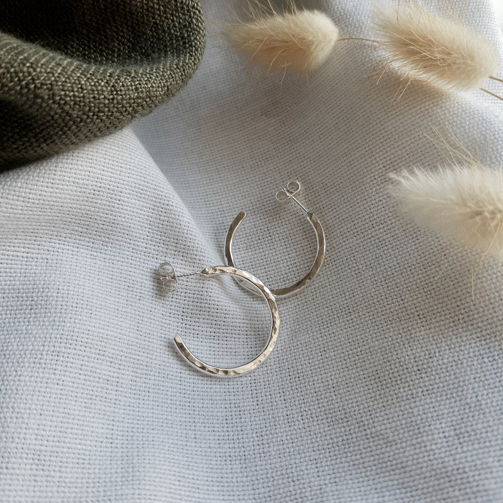 A pair of sterling silver hammered hoop earrings resting on a beige fabric surface with a fluffy white plant in the background.