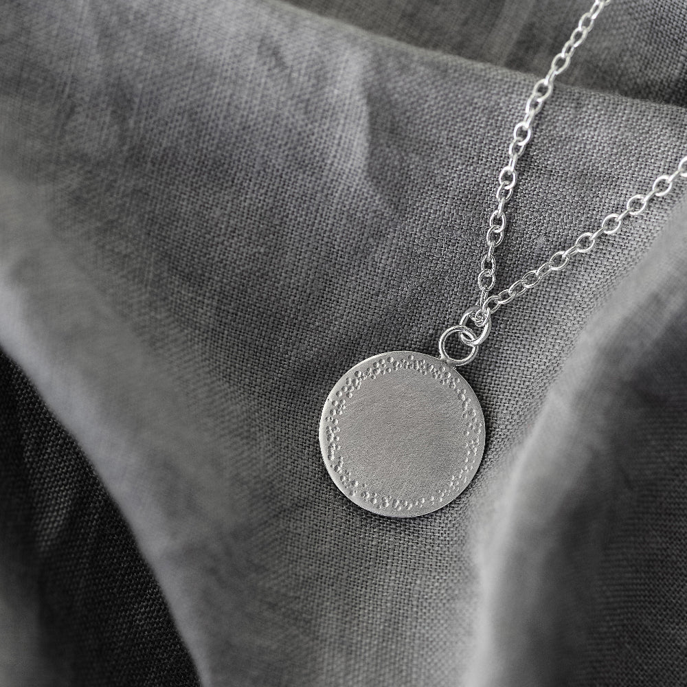 s silver pendant with a textured edge laying flat on a grey fabric background