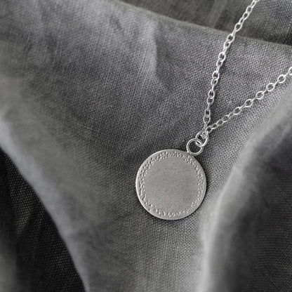 s silver pendant with a textured edge laying flat on a grey fabric background