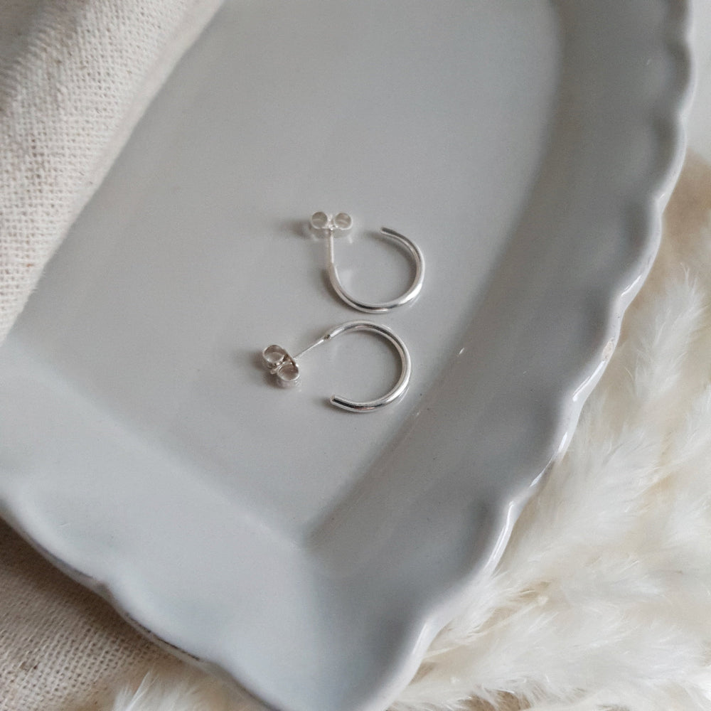 A pair of small sterling silver hoop earrings displayed on a white plate with a fuzzy white surface behind them.