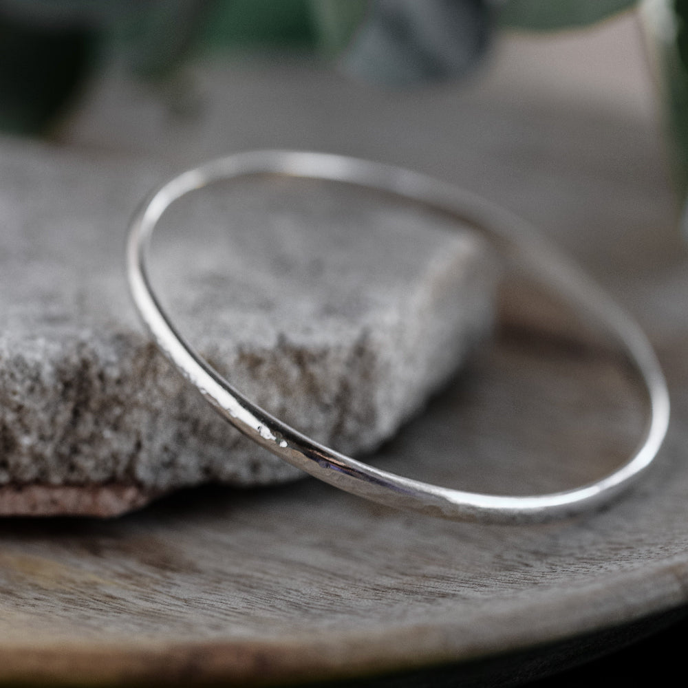 A silver bangle bracelet with a hammered texture, displayed on a grey surface.