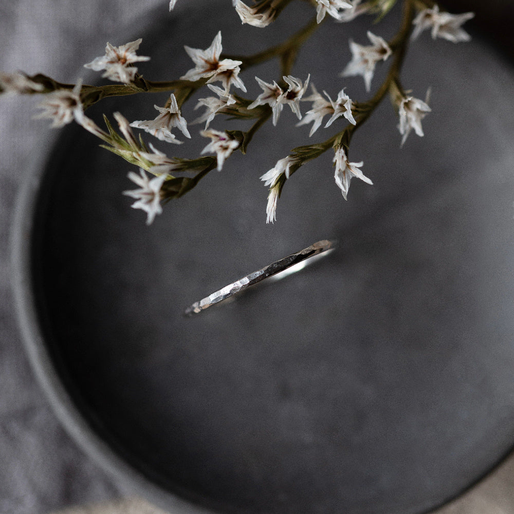 A single hammered silver stacking ring placed on a grey surface, with delicate white flowers hanging over it.