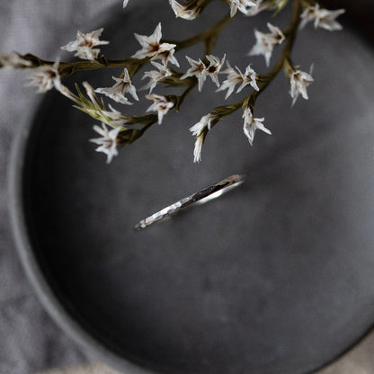 A single hammered silver stacking ring placed on a grey surface, with delicate white flowers hanging over it.