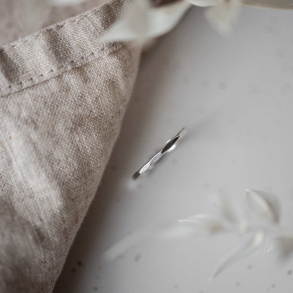 A simple, elegant silver stacking ring with a hammered finish, displayed on a neutral background with dried plants beside it.